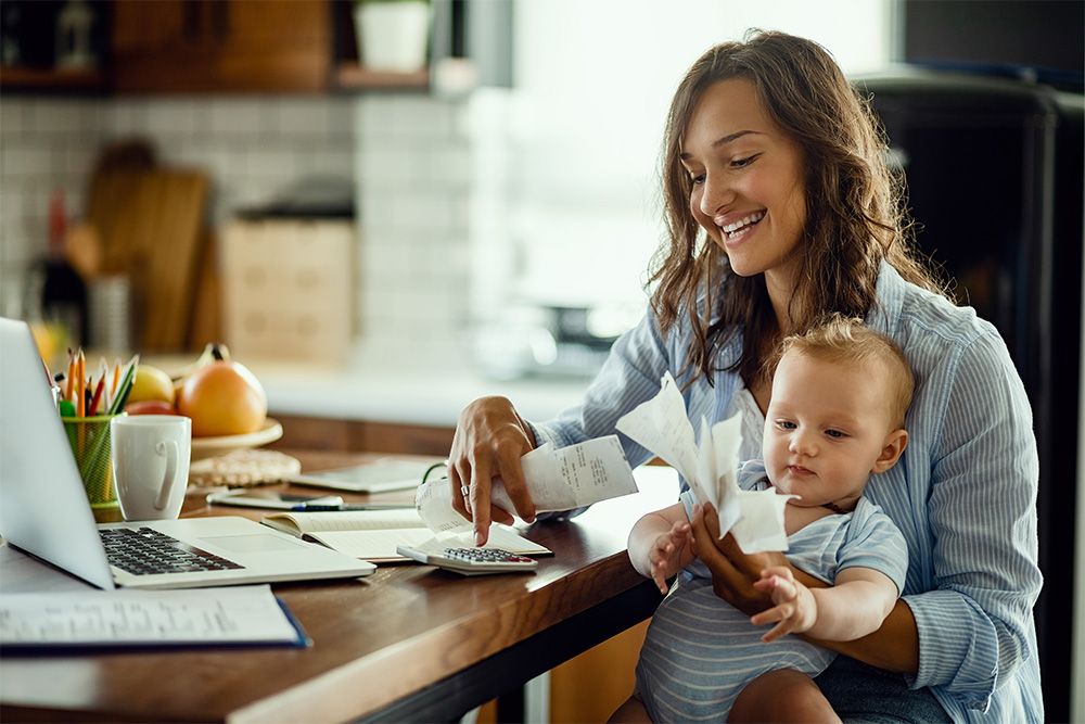 A woman and her baby sit at a table budgeting. There is a laptop and calculator on the table.