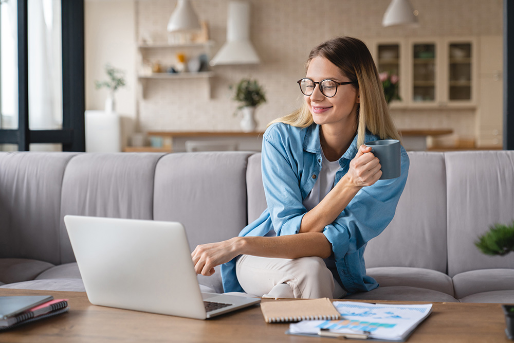 A young woman wearing glasses smiles down at a laptop computer as she sits on a couch and holds a coffee mug in her hand. 
