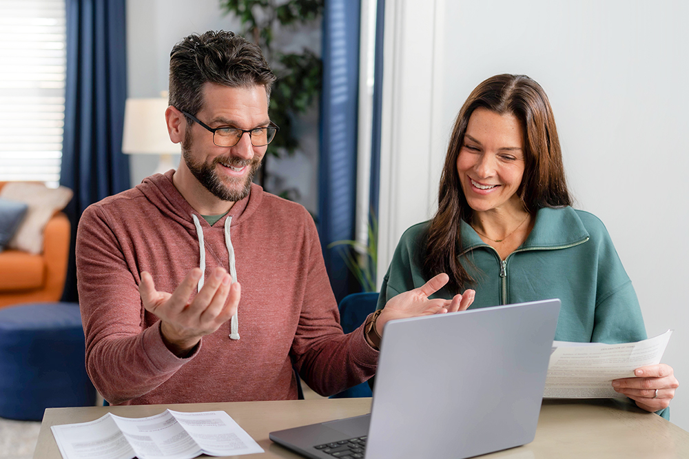 A man and woman sit at a table, looking at a laptop screen. They are smiling