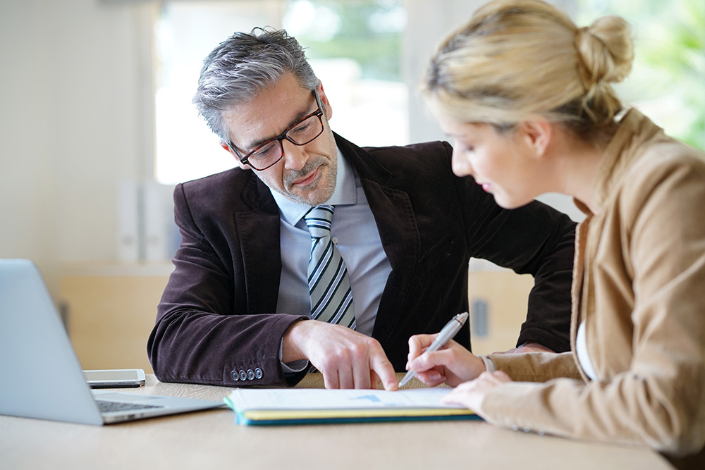 Man and woman sit at a table as the woman signs an official document for the pointing man.