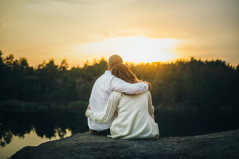 A man and woman sit on a rock watching the sunset. They each have one arm around the other in a hug.