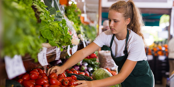 teen girl in apron stocking vegetables in supermarket