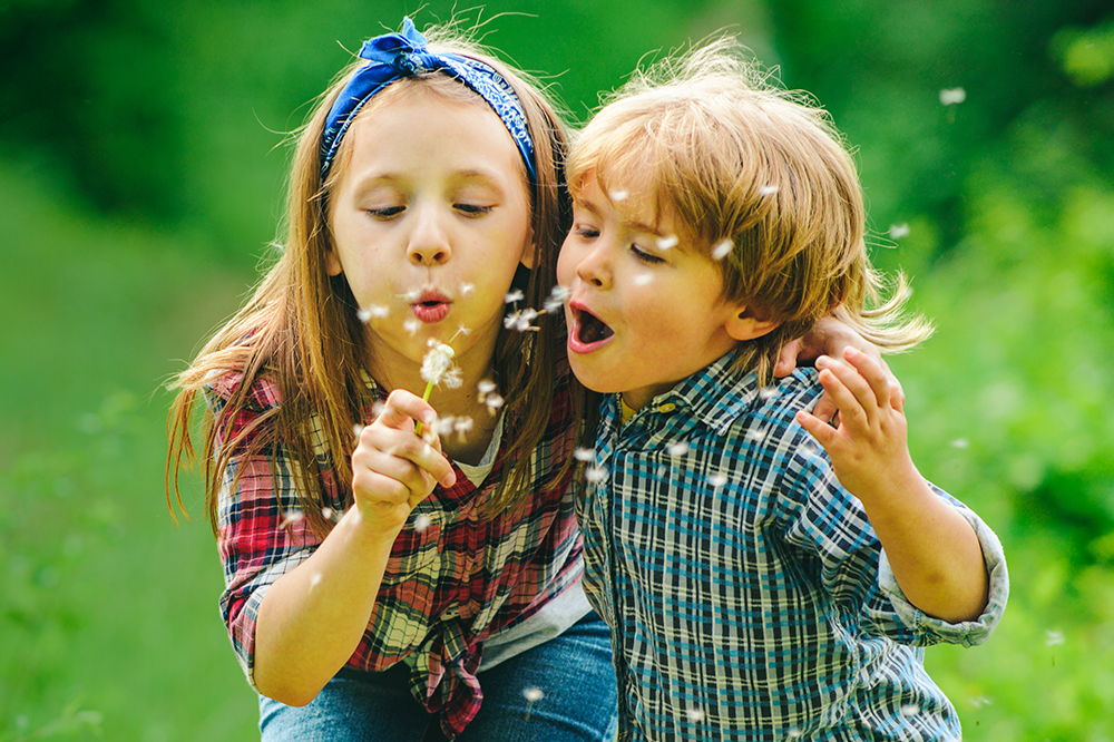A little girl and little blow blow a dandelion's seeds in the breeze outside.