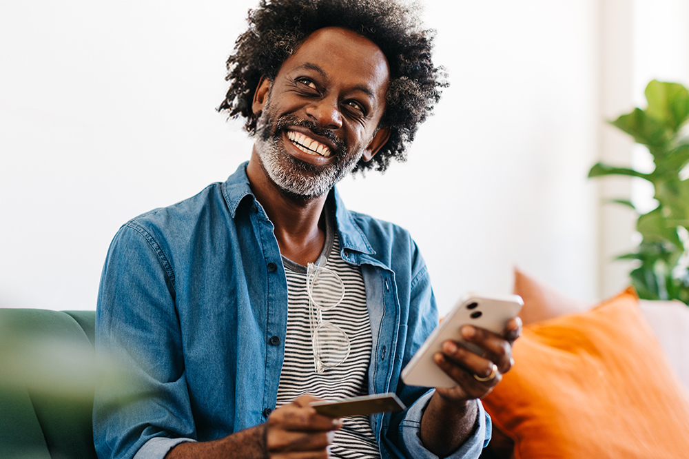 A man smiles as he looks out of frame, holding a smartphone in one hand and a credit card in the other. 