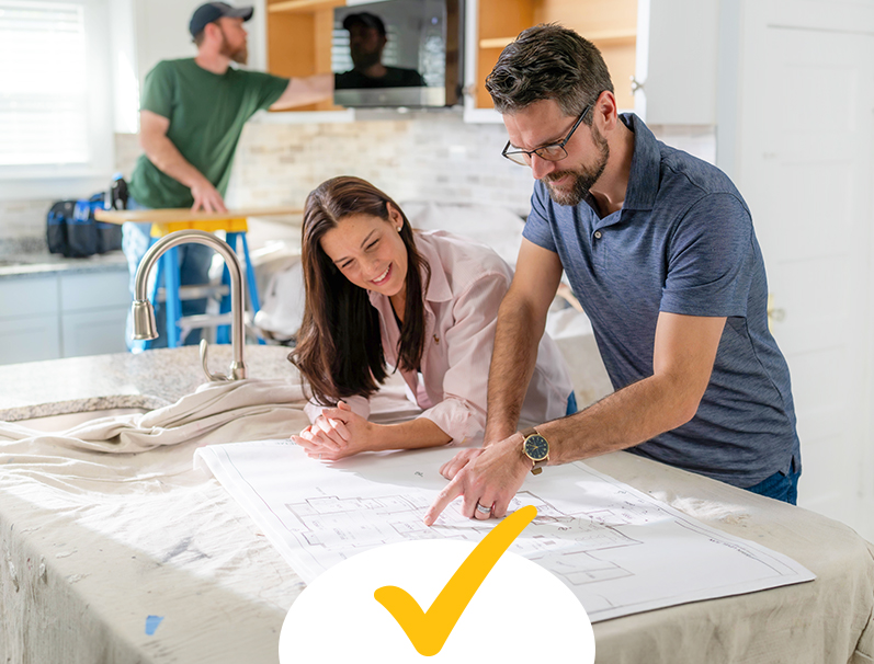 A couple leaning over a kitchen counter looking at blueprints while a handyman is on a ladder in the background