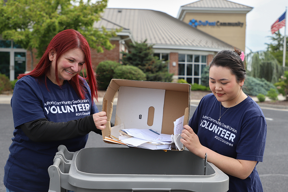 Two women in volunteer shirts dumping paper into a shred bin, standing in front of a credit union branch
